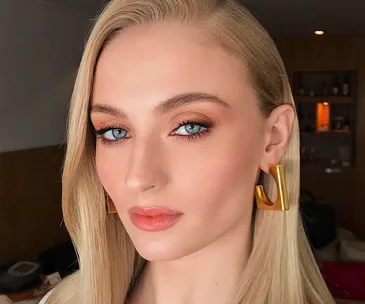 Woman with long blonde hair, wearing gold earrings, looking at the camera.