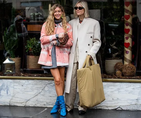 Two fashionable women in winter outfits; one in a pink jacket and blue boots, the other in a beige coat holding a large bag.