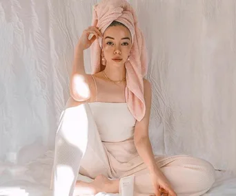 A woman sits cross-legged, wearing a pink towel on her head and cream loungewear, in a softly lit, minimalist setting.