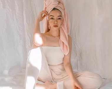 A woman sits cross-legged, wearing a pink towel on her head and cream loungewear, in a softly lit, minimalist setting.