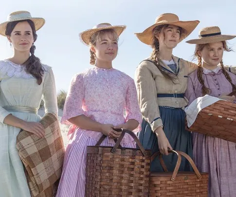 Four women in period dresses and hats hold picnic baskets, standing side by side outdoors.