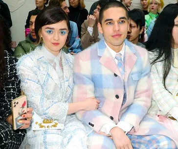 A couple in matching pastel plaid outfits sits at a fashion event, surrounded by an audience.