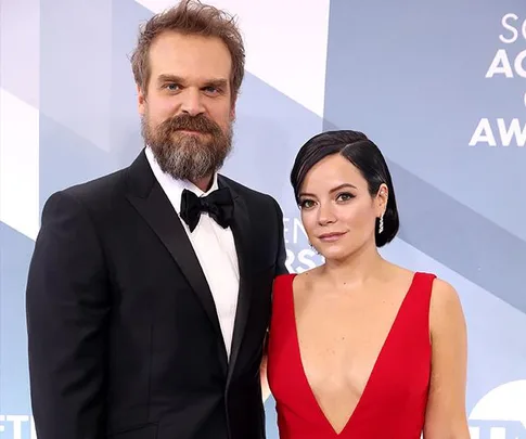 A man in a tuxedo and a woman in a red dress pose at a formal event with a blue and white backdrop.