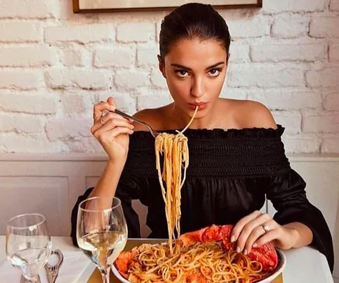 Woman in a black top eating spaghetti and lobster at a restaurant with white brick walls.