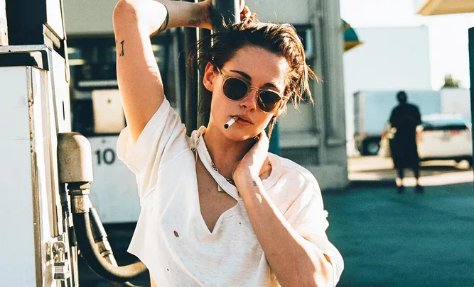 Person in sunglasses and white shirt at a gas station with a cigarette.