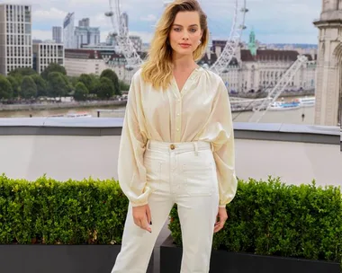 Woman in white blouse and pants stands on a rooftop with cityscape and Ferris wheel in the background.