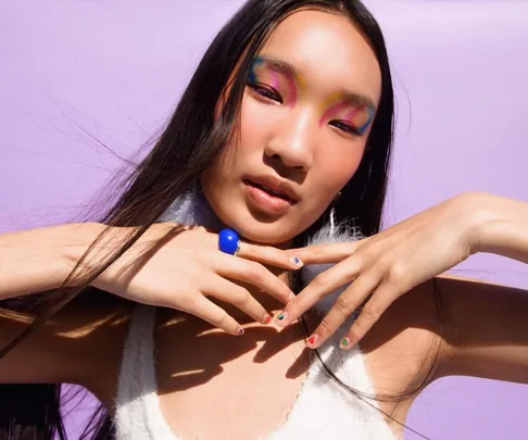 Person with colorful eye makeup and nail stickers posing against a purple background.