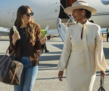 Two women smiling by a private jet, one in casual attire holding drinks, the other in an elegant dress and hat.