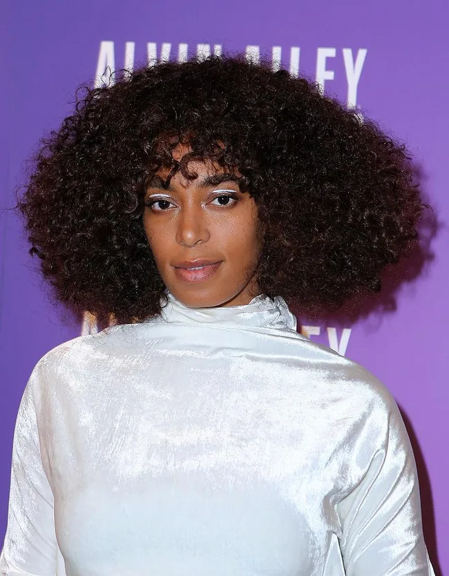 A person with curly hair wearing a shiny white top stands against a purple background.
