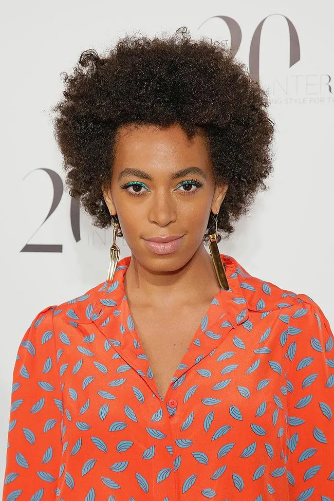 A person with an afro hairstyle wearing a patterned red blouse and long earrings against a white background.