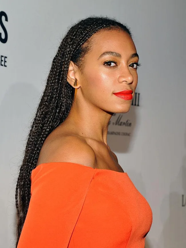 Woman in orange dress with braided hair and bold red lipstick, looking over shoulder at an event.