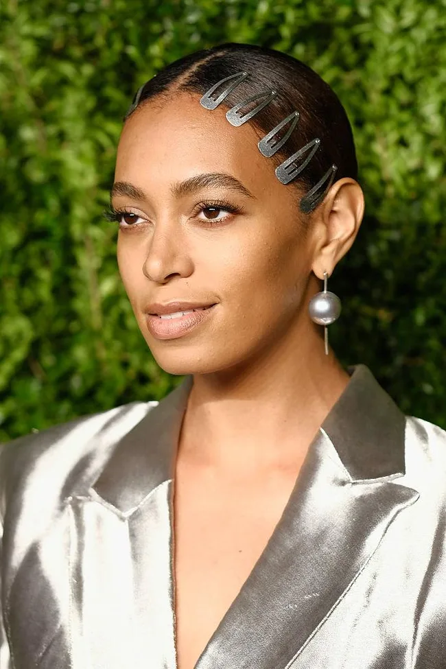 A person with slicked-back hair and metallic clips wears a silver outfit and pearl earring, standing in front of greenery.