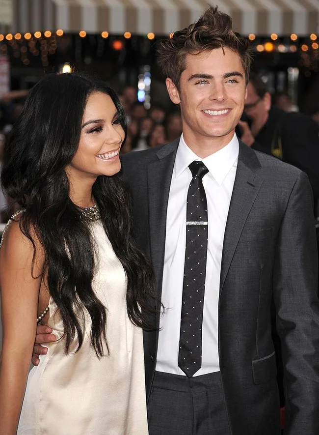 Young woman in a white dress and a man in a suit smile, posing together at an event.
