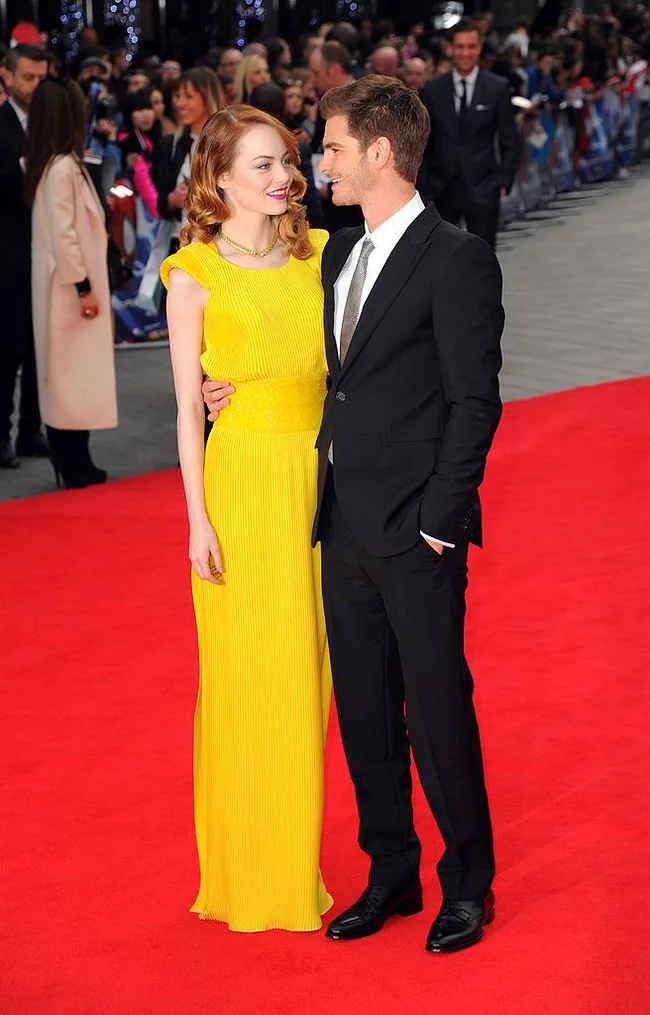 A woman in a yellow dress and a man in a suit smile at each other on a red carpet event.