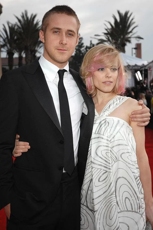 A man in a black suit and tie with a woman in a white dress with pink hair on a red carpet.