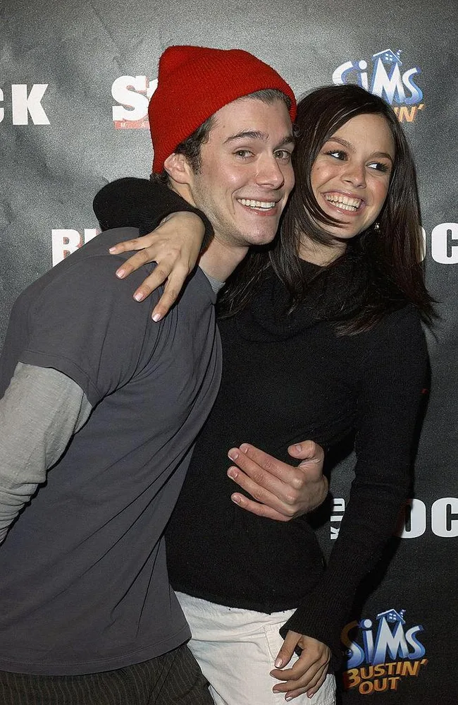 Two people smiling and embracing each other at a Sims Bustin' Out event, with a branded backdrop behind them.