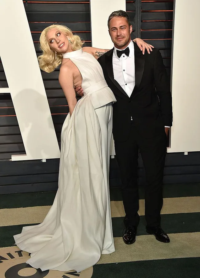Two people pose on the red carpet, one in a white dress and the other in a black tuxedo at a formal event.