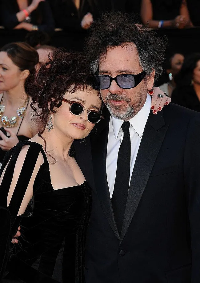 Couple in formal attire at an event, embracing and wearing sunglasses, with people blurred in the background.
