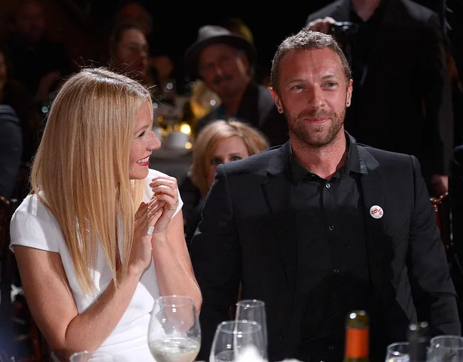 A woman in a white dress smiles at a man in a black suit at a formal event, seated at a table with glasses and bottles.