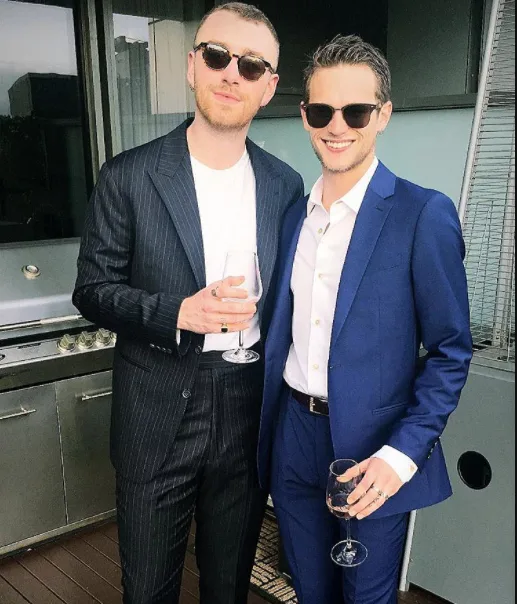 Two people in sunglasses and suits, holding wine glasses, standing near an outdoor grill, smiling at the camera.