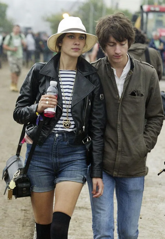 A woman in a hat, striped shirt, and leather jacket with a man in a green jacket walk on a dirt path with festivalgoers.