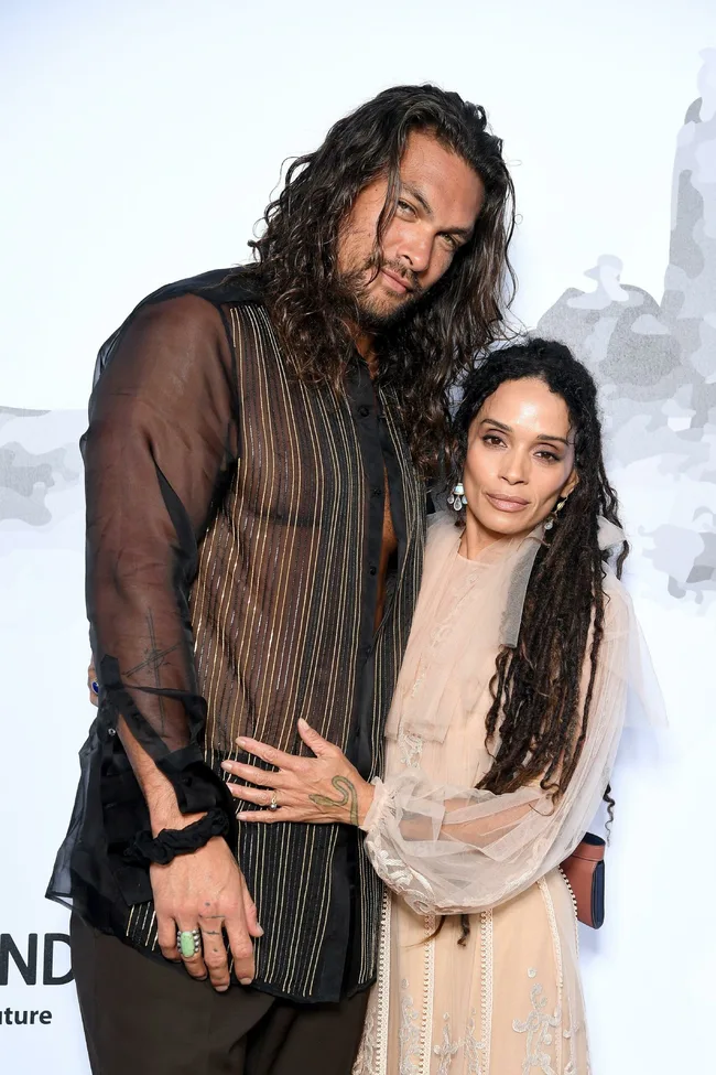 Man in a sheer black shirt with striped design poses with a woman in a lace dress, both smiling at the camera.