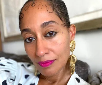 Woman with large gold earrings and pink lipstick, wearing a polka-dotted shirt, looking directly at the camera.