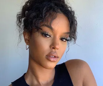 Person with curly hair, wearing a black top and hoop earrings, posing against a light background.