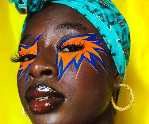 African woman with vibrant face paint, blue headwrap, and gold hoop earrings against a yellow background.