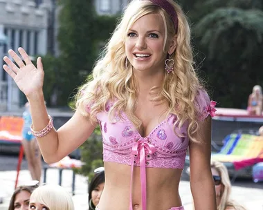 A woman with blonde hair waves, wearing a pink crop top and accessories, smiling outdoors with blurred people in the background.