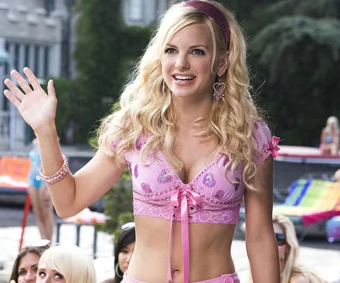 A woman with blonde hair waves, wearing a pink crop top and accessories, smiling outdoors with blurred people in the background.