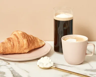 A croissant on a plate, a glass of coffee, a cup of coffee, and a spoon with collagen powder on a marble surface.