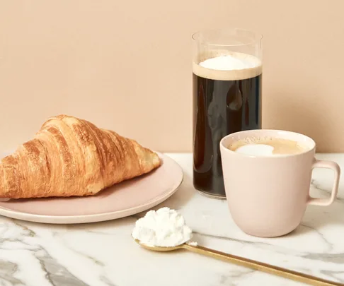 A croissant on a plate, a glass of coffee, a cup of coffee, and a spoon with collagen powder on a marble surface.