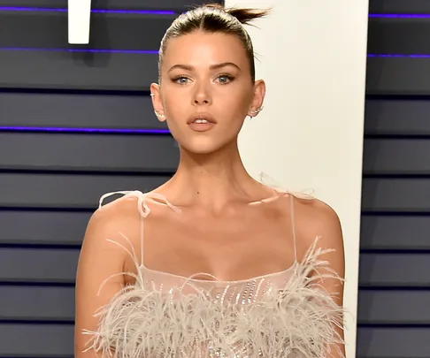 Model with sleek bun hairstyle, wearing elegant feathered dress, posing at an event with neutral makeup.