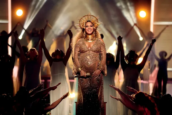 Beyoncé stands in a gold gown and headdress, surrounded by dancers with arms raised in a dynamic stage performance.