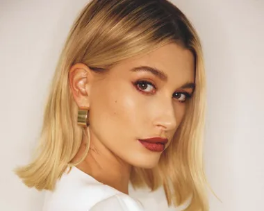 Blonde woman with side-parted bob, wearing red lipstick and gold earrings, gazing past the camera.