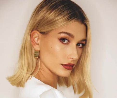 Blonde woman with side-parted bob, wearing red lipstick and gold earrings, gazing past the camera.