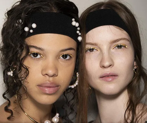 Two models wearing black headbands with pearls, showcasing natural makeup with gold eyeliner.