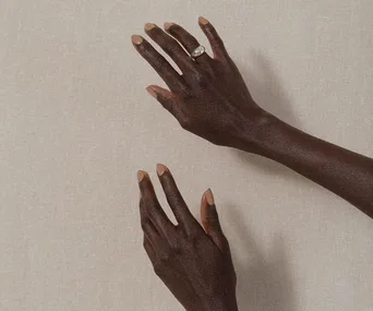 Two hands with nude nail polish on a beige background, one wearing a silver ring.