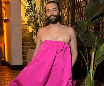 Jonathan Van Ness wearing a vibrant pink dress, standing in an elegant interior with plants.