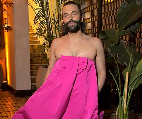 Jonathan Van Ness wearing a vibrant pink dress, standing in an elegant interior with plants.