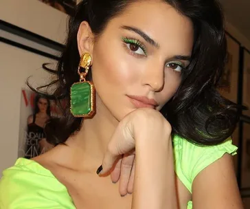 Person with bold green eye makeup and large green earrings, wearing a bright green top, posing thoughtfully indoors.