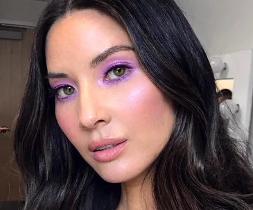 Woman with long dark hair wearing pastel purple eyeshadow, soft pink blush, and nude lipstick, close-up portrait.