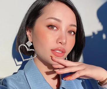 Woman with short black hair, wearing large heart-shaped earrings, rests hand on chin, in soft natural light.