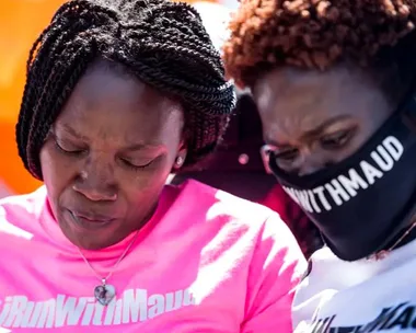 Two people wearing "IRunWithMaud" shirts, one with a mask, stand in a solemn pose.