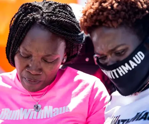 Two people wearing "IRunWithMaud" shirts, one with a mask, stand in a solemn pose.