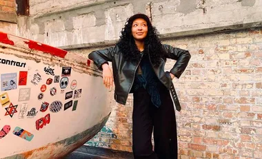 A person with curly hair and a leather jacket stands by a sticker-covered wall in an industrial setting.