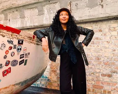 A person with curly hair and a leather jacket stands by a sticker-covered wall in an industrial setting.