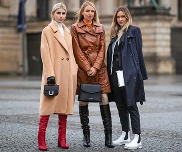 Three women wearing stylish winter coats and boots, posing confidently on a city street.