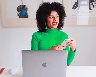 Person in a bright green sweater smiling, holding a phone, sitting at a desk with a laptop.